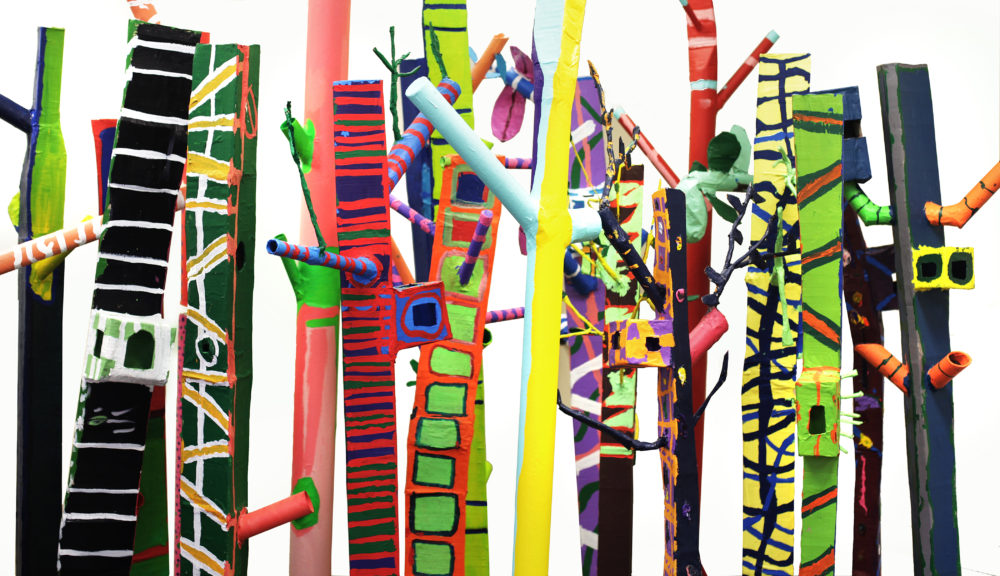 This image features a colorful and intricate installation of painted wooden poles. The poles are arranged closely together, creating a dense forest-like effect.