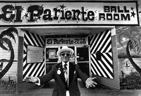 Man in suit with open arms in front of a ball room, black and white photo, "Americanos: Latino Life in the United States"