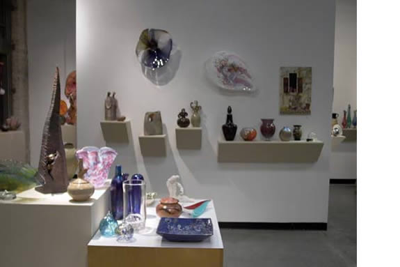 On the shelves along the wall, there is a wide variety of artwork, including: Glass sculptures: These are particularly striking, with intricate shapes and vibrant colors. Ceramics: There are several ceramic pieces, ranging from functional vases to more sculptural forms. Paintings: Smaller paintings in different styles are displayed on the shelves. On the floor, a display case showcases a collection of glass and ceramic objects, including vases, bowls, and figurines.