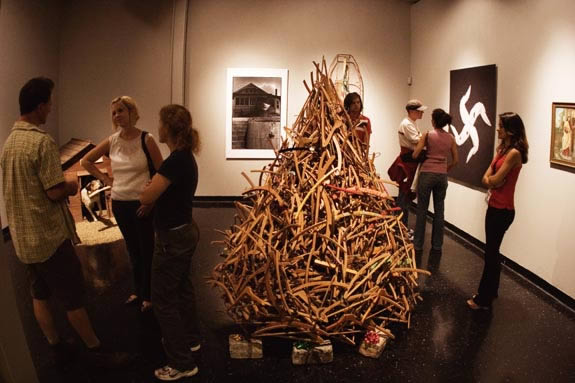 People converse in a gallery space. In the center is a tree shaped structure made out of gathered wood.