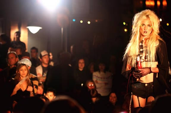 Blonde woman in foreground on a stage with figures dimly lit behind her