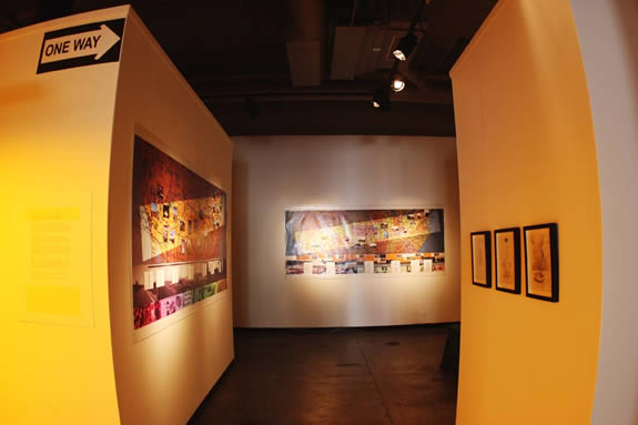 An interior view of an art gallery with a "ONE WAY" sign prominently displayed. A large, colorful mural covers one wall, depicting a scene with multiple figures and objects. Smaller framed artworks are displayed on another wall.