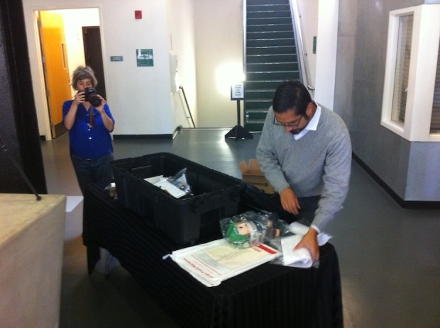 Woman in background photographs man in foreground as he packs objects on a black cloth covered table.