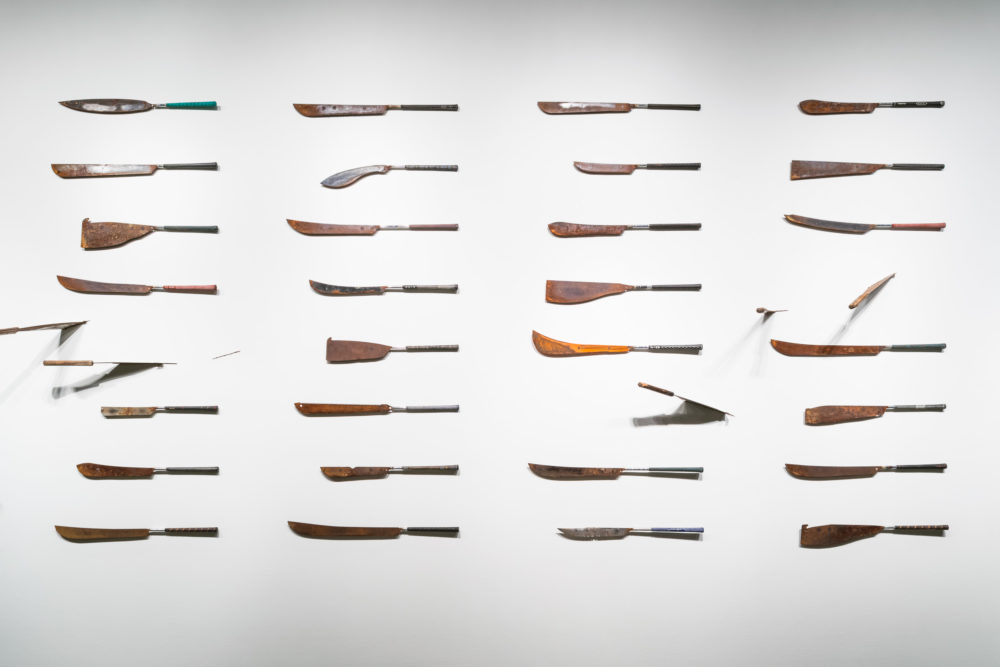 The weapons are arranged in a grid pattern, each one mounted on a separate peg. They appear to be various types of knives or machetes, with different blade shapes, sizes, and materials. Some have wooden handles, while others have metal ones. The blades range from smooth and polished to textured and rough.