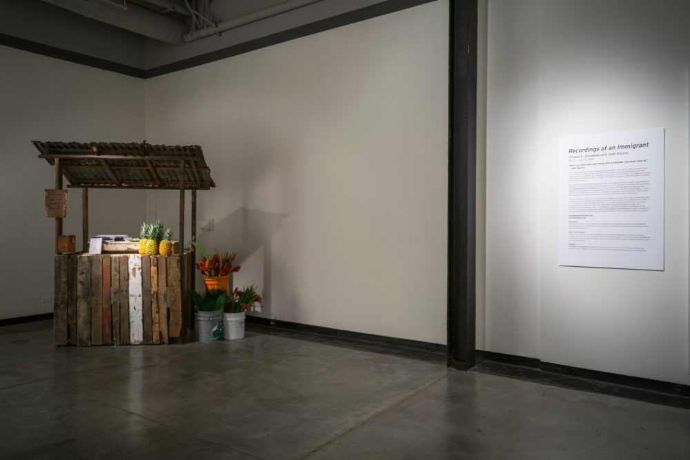 Small wooden stall with fruits and flowers. On the right is the wall text for the exhibition.