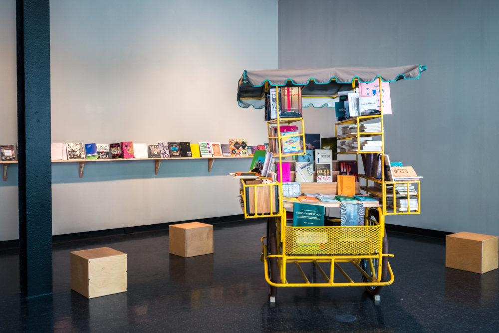 The bookmobile is a bright yellow cart with a canvas canopy, resembling a miniature bookstore on wheels.