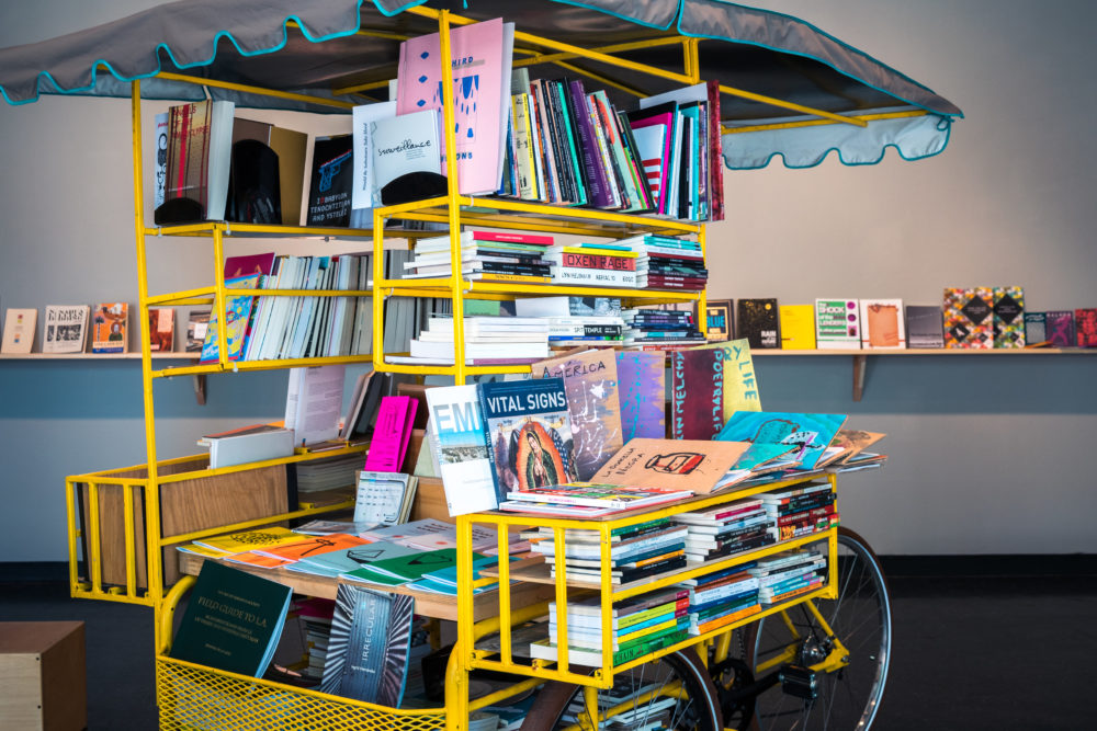 The bookmobile is a bright yellow cart with a canvas canopy, resembling a miniature bookstore on wheels.