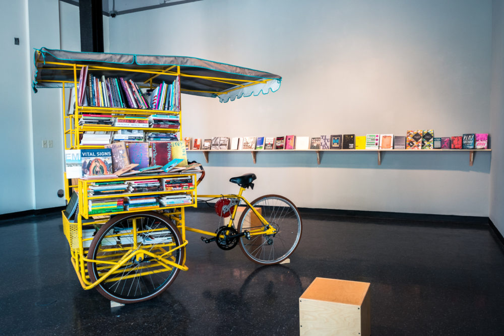 The bookmobile is a bright yellow cart with a canvas canopy, resembling a miniature bookstore on wheels.