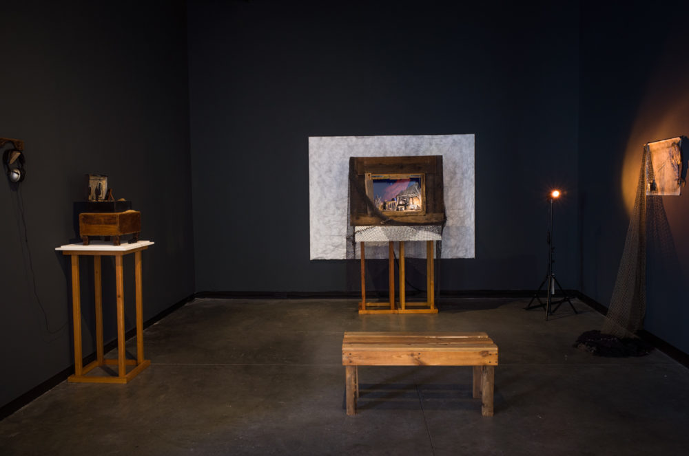 The gallery space is dimly lit, with a dark gray or black wall dominating the background. In the center of the wall, a rectangular frame stands out, painted white and bordered by a wooden frame. Within the frame, a small, seemingly hand-crafted scene is visible, illuminated by a spotlight.