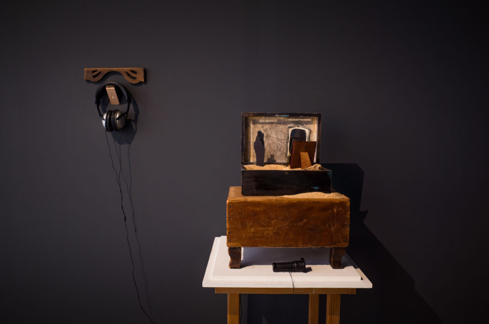 In a darkened room, a vintage wooden box sits atop a white pedestal. The box is open, revealing a miniature scene inside, illuminated by a small light source. The scene appears to be a miniature theater stage, with tiny figures and props.