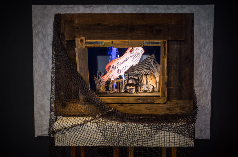 The diorama is set within a wooden frame, reminiscent of a picture window. Inside, a miniature scene is illuminated, depicting a fisherman