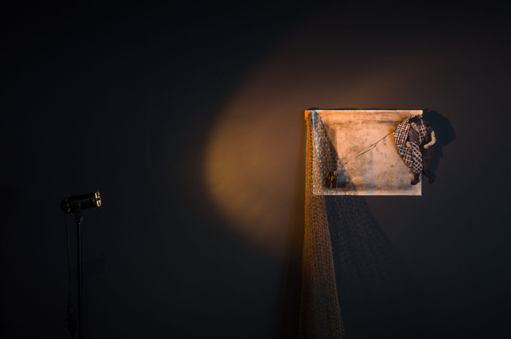 In a darkened room, a spotlight illuminates a rectangular canvas mounted on the wall. The canvas depicts a simple scene: a plaid shirt draped over a fishing net, with a small object, possibly a fishing lure, attached. The scene is starkly lit, casting long shadows on the wall.