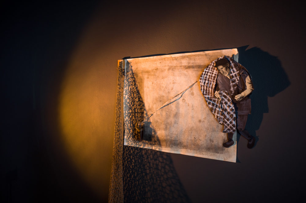 A rectangular canvas is mounted on a dark wall, illuminated by a spotlight. The canvas appears to be a weathered piece of paper with a fishing net draped over it. A small, lifelike figure of a man is positioned on the paper, seemingly caught in the net.