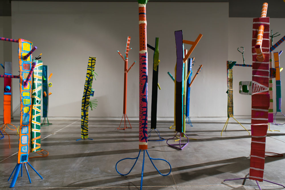 The sculptures are made of various materials, including pipes, strips of fabric, and other found objects. They are painted in a variety of bright colors, including red, yellow, blue, and green. The sculptures are arranged in a seemingly random pattern throughout the gallery space, casting long shadows on the floor.