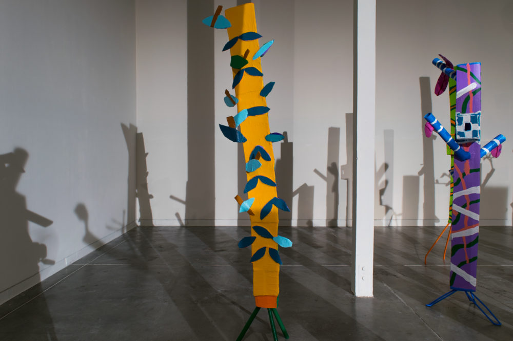 The sculptures are made of various materials, including pipes, strips of fabric, and other found objects. They are painted in a variety of bright colors, including red, yellow, blue, and green. The sculptures are arranged in a seemingly random pattern throughout the gallery space, casting long shadows on the floor.