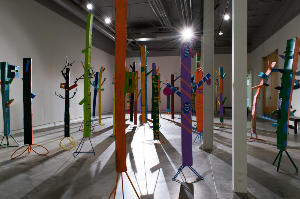 The sculptures are made of various materials, including pipes, strips of fabric, and other found objects. They are painted in a variety of bright colors, including red, yellow, blue, and green. The sculptures are arranged in a seemingly random pattern throughout the gallery space, casting long shadows on the floor.