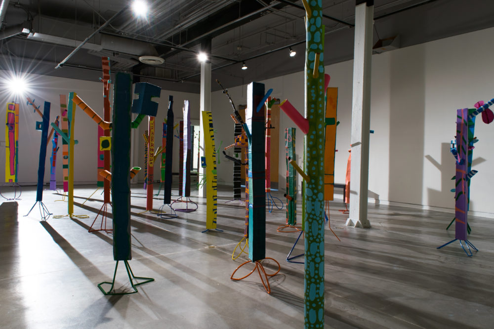 The sculptures are made of various materials, including pipes, strips of fabric, and other found objects. They are painted in a variety of bright colors, including red, yellow, blue, and green. The sculptures are arranged in a seemingly random pattern throughout the gallery space, casting long shadows on the floor.