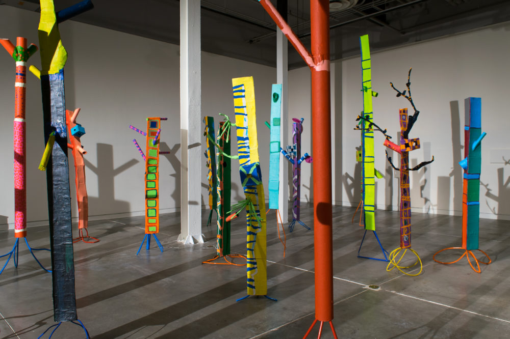 The sculptures are made of various materials, including pipes, strips of fabric, and other found objects. They are painted in a variety of bright colors, including red, yellow, blue, and green. The sculptures are arranged in a seemingly random pattern throughout the gallery space, casting long shadows on the floor.