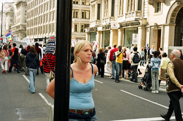 Blond woman standing next to post with protest occurring on road behind her.