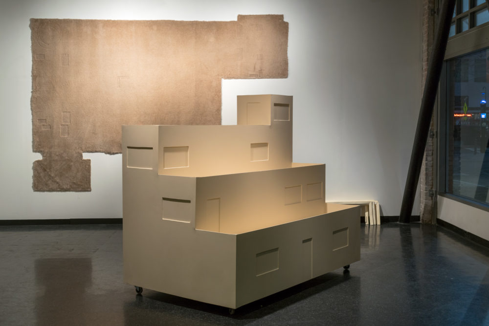 This image depicts a contemporary art installation in a gallery setting. The central focus is a large, multi-tiered cardboard structure resembling a miniature building or architectural model. It