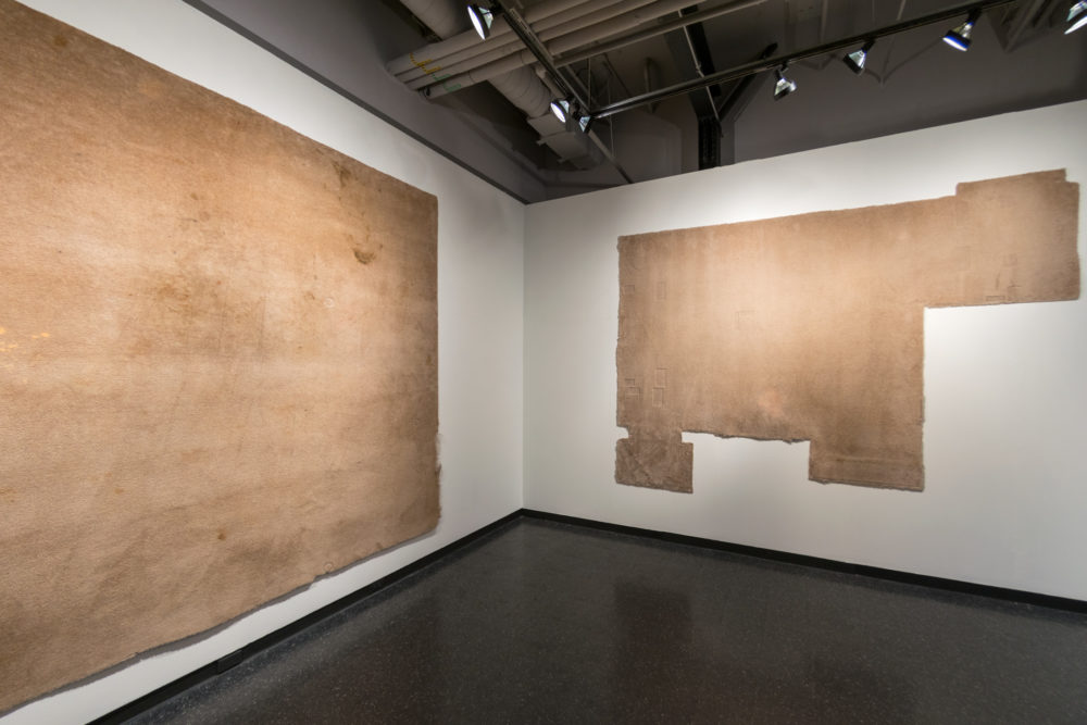This image depicts a contemporary art installation in a gallery setting. The focus is on two large, rectangular pieces of textile or fabric hanging on opposite walls. Both pieces are a muted brown color and have a rough, textured surface. The one on the left is larger and more intact, while the one on the right is smaller and appears to be partially damaged or incomplete.