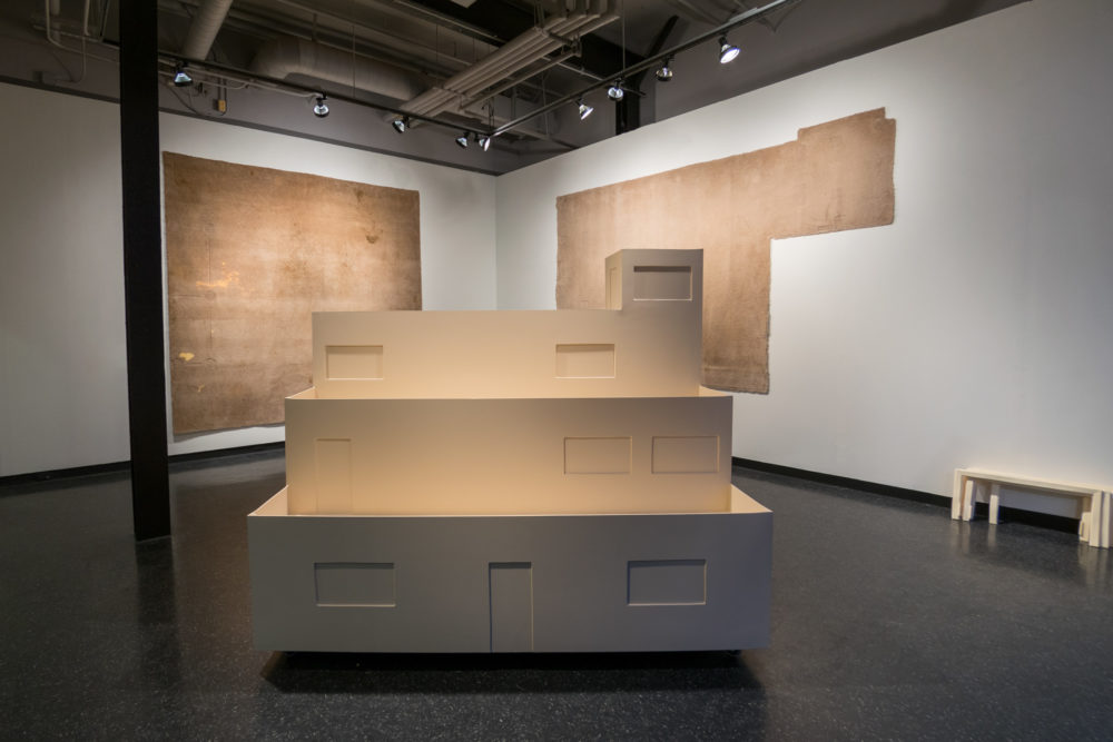 The central focus is a large, multi-tiered cardboard structure resembling a miniature building or architectural model. It