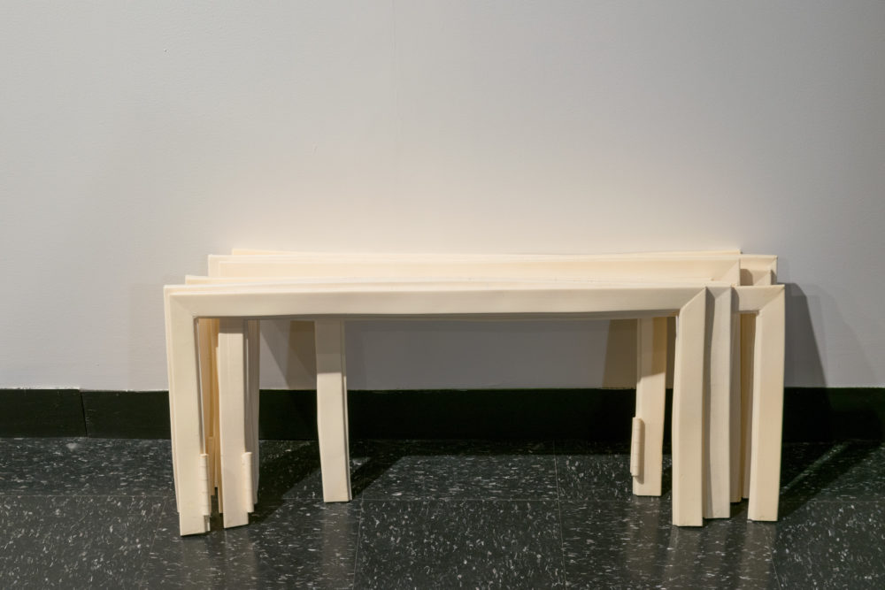 This image shows a close-up of a group of rectangular frames stacked on top of each other. The frames are made of a white or off-white material, possibly ceramic or porcelain. They are arranged in a leaning position, with one end resting on the floor and the other leaning against a wall. Some of the frames are slightly tilted or offset.