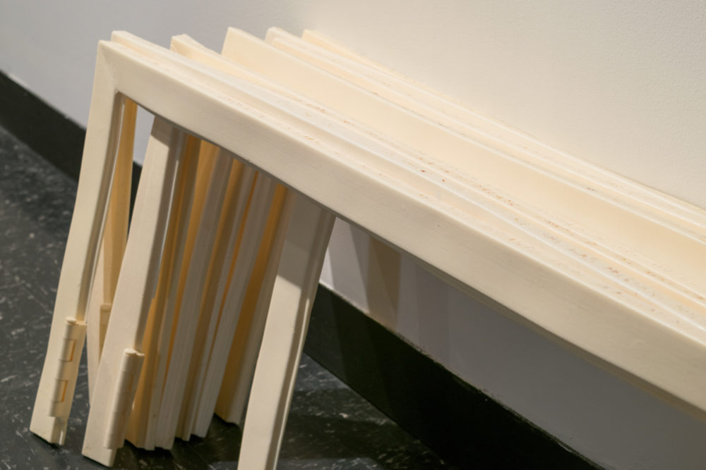This image shows a close-up of a group of rectangular frames stacked on top of each other. The frames are made of a white or off-white material, possibly ceramic or porcelain. They are arranged in a leaning position, with one end resting on the floor and the other leaning against a wall. Some of the frames are slightly tilted or offset.