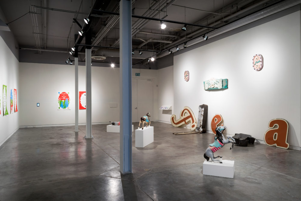 Several large, vintage neon signs are scattered throughout the gallery. Some are intact, while others are broken or partially disassembled. The signs feature various colors and designs, including letters, symbols, and abstract shapes. A brightly painted carousel horse stands on a pedestal, adding a whimsical element to the exhibition. Smaller pieces of artwork, such as paintings and sculptures, are also on display on the walls.