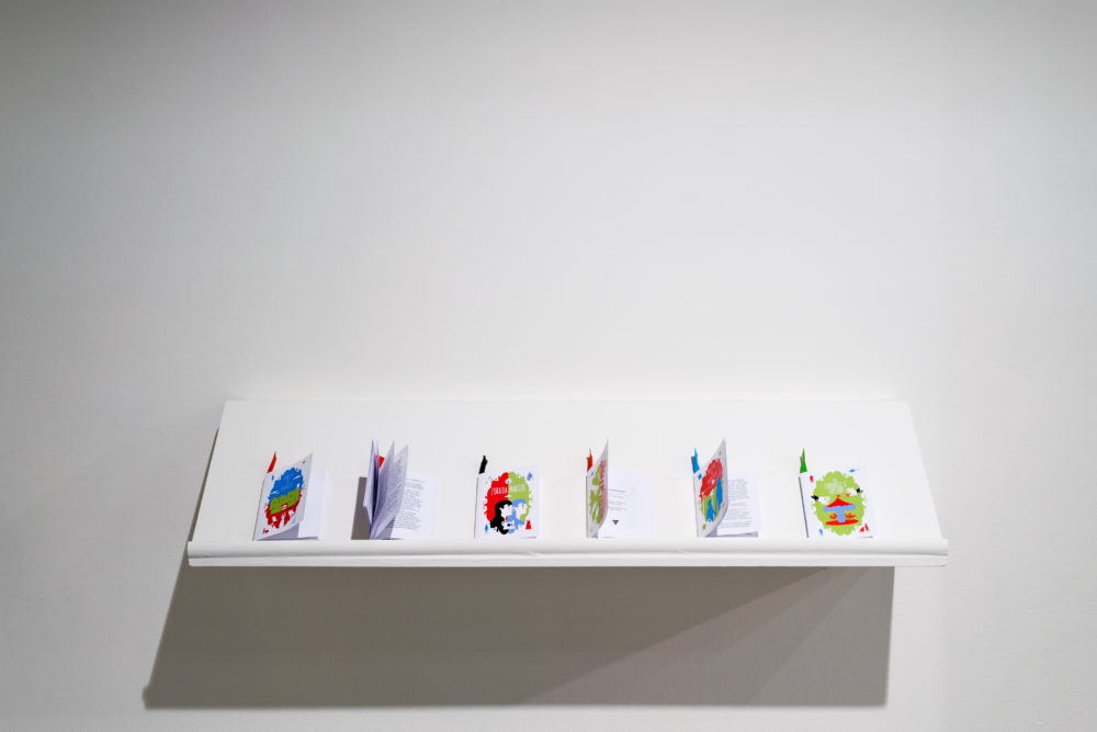 A clean, white shelf extends horizontally from the wall. Several small, colorful booklets are arranged on the shelf. They are folded and have different sizes and designs.