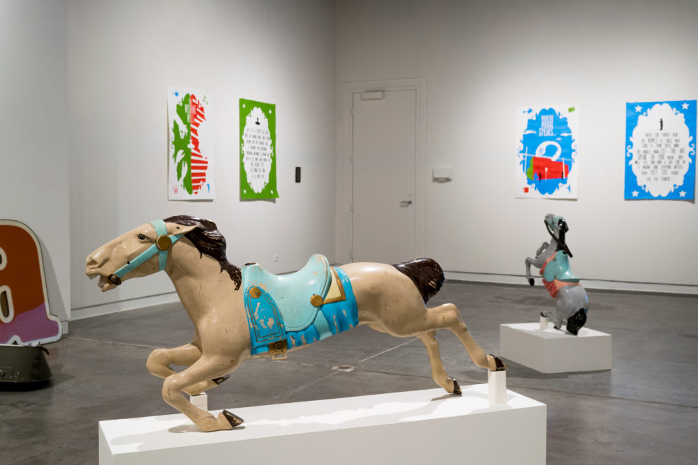 Two brightly painted carousel horses are prominently displayed on pedestals. One is a classic white horse with a blue saddle, while the other is a smaller, gray horse with a more abstract design.Several colorful posters with playful designs and text are hung on the walls. They add a sense of energy and vibrancy to the space.