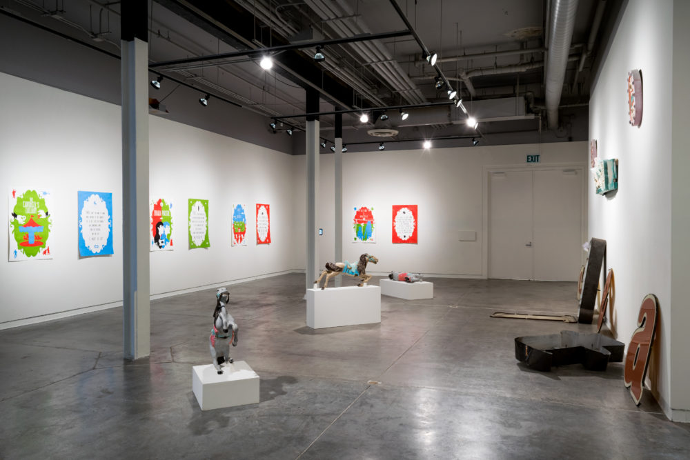 Several large, vintage neon signs are scattered throughout the gallery. Some are intact, while others are broken or partially disassembled. The signs feature various colors and designs, including letters, symbols, and abstract shapes. A brightly painted carousel horse stands on a pedestal, adding a whimsical element to the exhibition. Smaller pieces of artwork, such as paintings and sculptures, are also on display on the walls.
