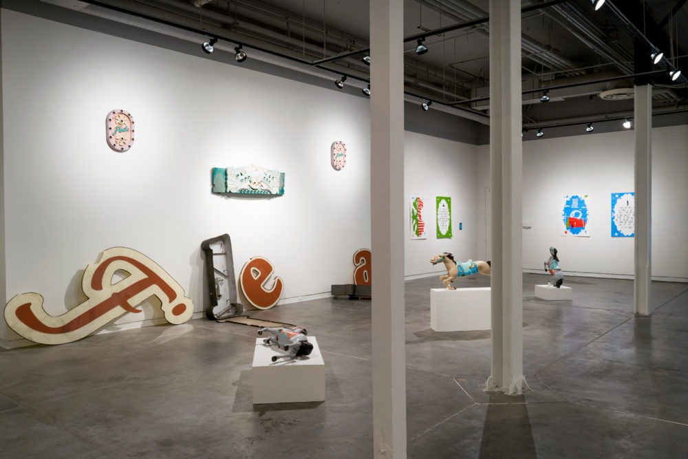 Several large, vintage neon signs are scattered throughout the gallery. Some are intact, while others are broken or partially disassembled. The signs feature various colors and designs, including letters, symbols, and abstract shapes. A brightly painted carousel horse stands on a pedestal, adding a whimsical element to the exhibition. Smaller pieces of artwork, such as paintings and sculptures, are also on display on the walls.