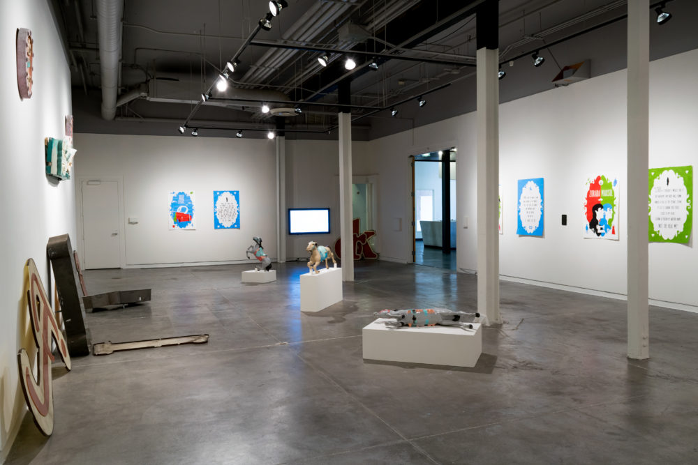 Several large, vintage neon signs are scattered throughout the gallery. Some are intact, while others are broken or partially disassembled. The signs feature various colors and designs, including letters, symbols, and abstract shapes. A brightly painted carousel horse stands on a pedestal, adding a whimsical element to the exhibition. Smaller pieces of artwork, such as paintings and sculptures, are also on display on the walls.