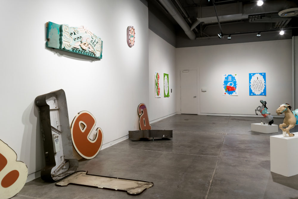 Several large, vintage neon signs are scattered throughout the gallery. Some are intact, while others are broken or partially disassembled. The signs feature various colors and designs, including letters, symbols, and abstract shapes. A brightly painted carousel horse stands on a pedestal, adding a whimsical element to the exhibition. Smaller pieces of artwork, such as paintings and sculptures, are also on display on the walls. These include a colorful painting of a face and a sculptural piece resembling a neon sign.