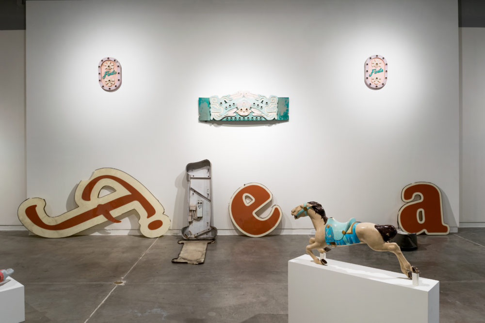 Several large, vintage neon signs are scattered throughout the gallery. Some are intact, while others are broken or partially disassembled. The signs feature various colors and designs, including letters, symbols, and abstract shapes. A brightly painted carousel horse stands on a pedestal, adding a whimsical element to the exhibition. Smaller pieces of artwork, such as paintings and sculptures, are also on display on the walls. These include a colorful painting of a face and a sculptural piece resembling a neon sign.
