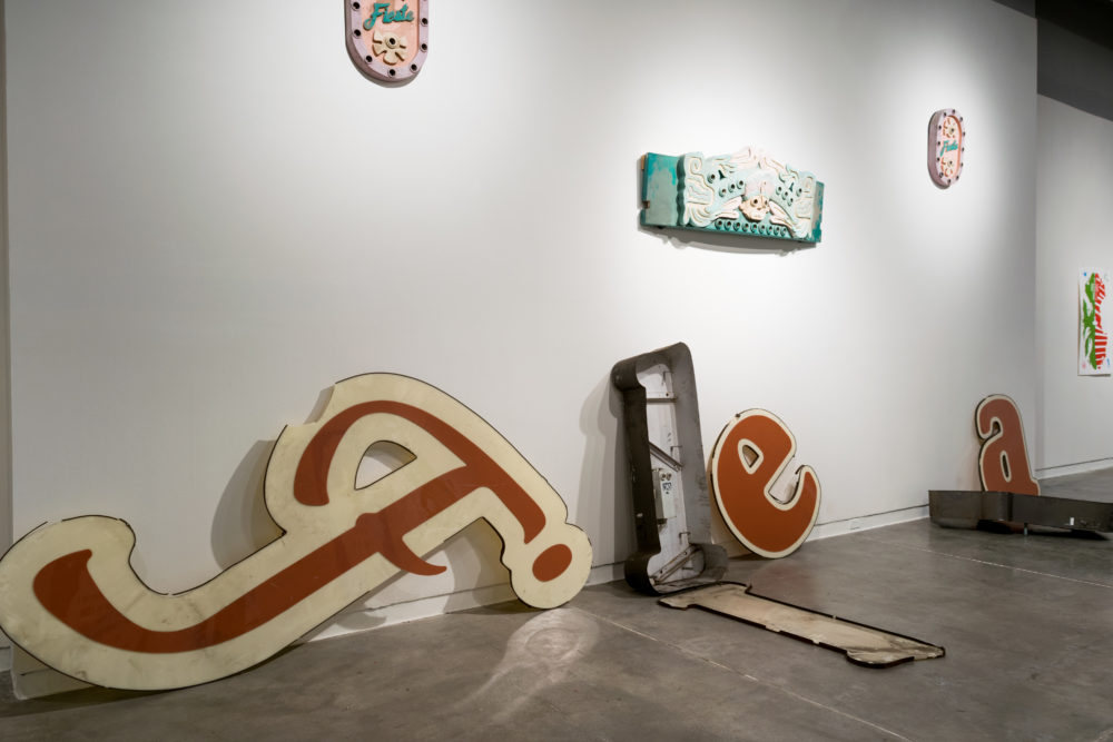 Several large, vintage signs are scattered throughout the gallery. Some are intact, while others are broken or partially disassembled. The signs feature various colors and designs, including letters, symbols, and abstract shapes. Smaller pieces of artwork, such as paintings and sculptures, are also on display on the walls.