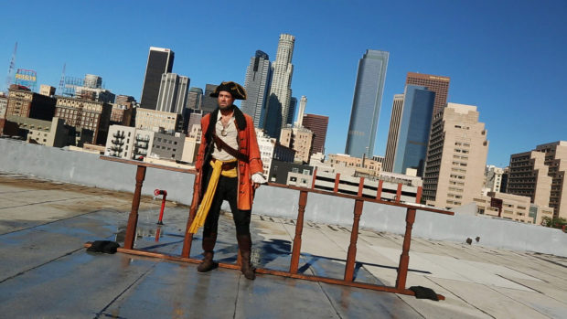 A male pirate stands against a railing on a rooftop with the city of Los Angeles in the background