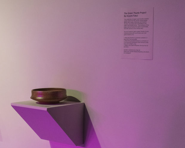 A white panel on the wall displays information about the artwork titled "The Green Thumb Project" by Kiyomi Fukui. The text describes the process of creating paper cast thumbs containing seeds and encourages viewers to take one home and plant it. A small, brown ceramic bowl is placed on a white shelf that protrudes from the wall. The bowl appears to be a simple, handmade piece.