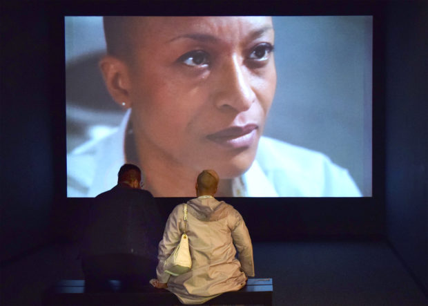 Two figures sitting on a bench, watching "Exchange," video installation