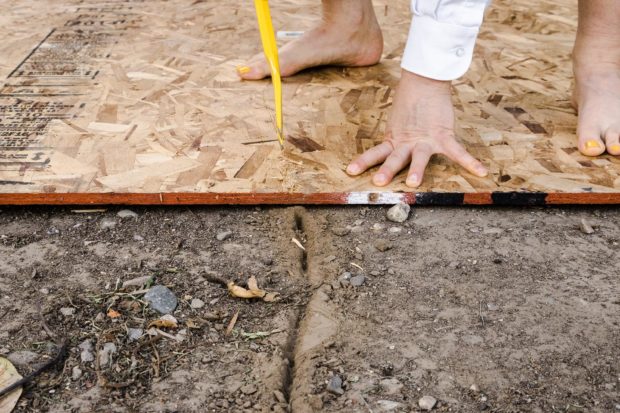 individual dressed in white uses a crowbar to cut ridge into dirt and board