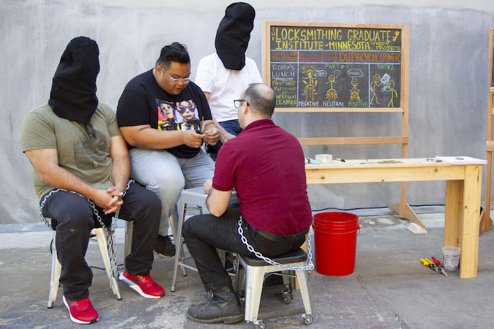 Artist seated with 3 participants, 2 are wearing black hoods
