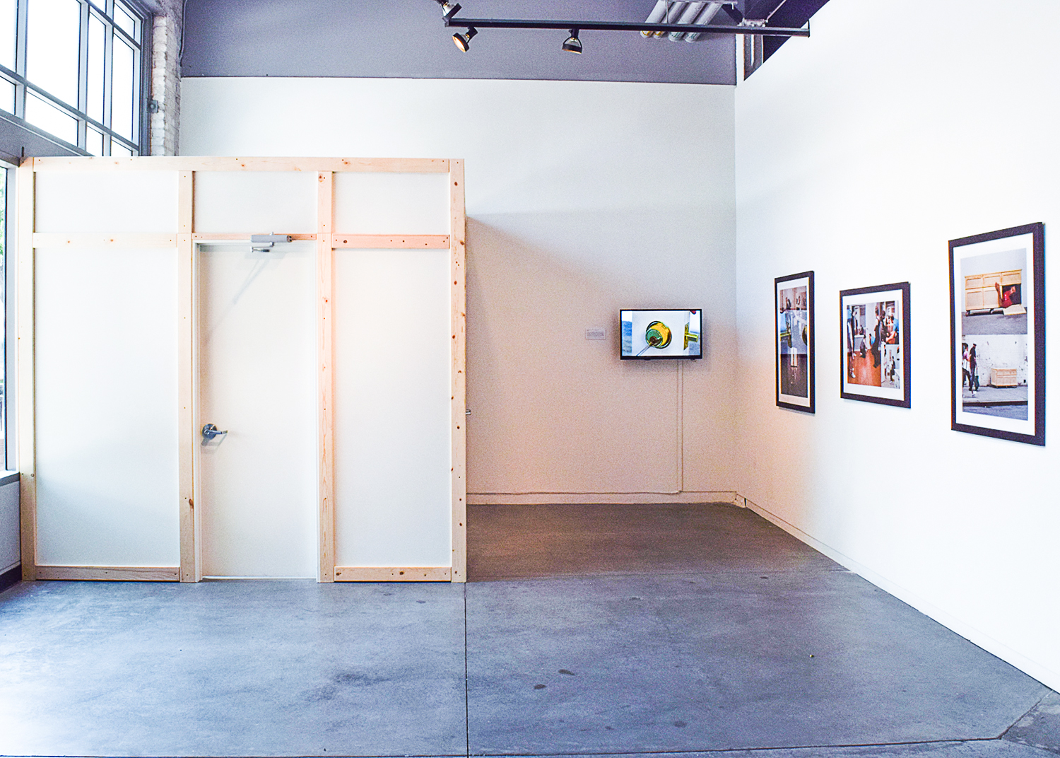 White Structure: A white, wooden-framed structure occupies a significant portion of the space. It has a door and appears to be a room within a room. Television: A television mounted on the wall within the structure displays a video or image. Framed Photographs: Three framed photographs hang on the wall opposite the structure, showcasing black and white images of people and possibly urban landscapes.