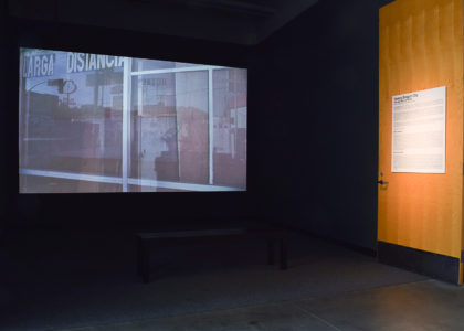 Projection Screen: A large projection screen dominates one wall, displaying a video or film. The content appears to be grainy and nostalgic, with images of a storefront and street scene. Sergio De La Torre Nuevo Dragon City