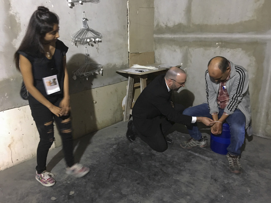 Artist teaching a man how to break out from zip ties while a young woman watches the performance.