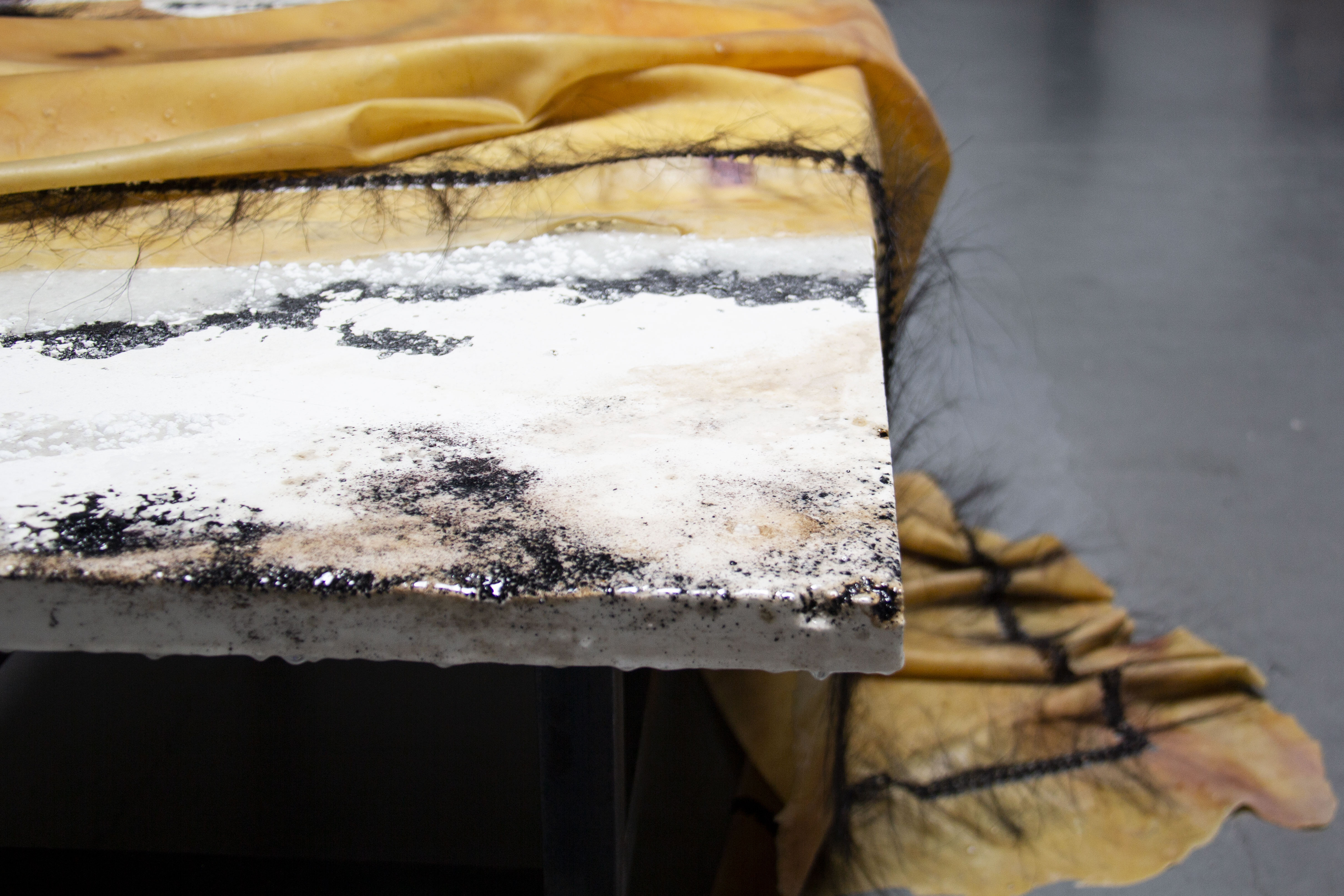 The image depicts a close-up of an abstract artwork. A rectangular canvas, partially covered by a yellow fabric, is the primary focus. The visible surface of the canvas is textured and layered with various materials, including: White Base: A white, possibly plaster-like base forms the foundation of the artwork. Dark Marks: Dark, almost charcoal-like marks are scattered across the white base, creating a sense of depth and texture. Yellow Fabric: A yellow, possibly silk or satin fabric drapes over the top edge of the canvas. It appears to be attached by a series of fine black threads. Other Textures: There are hints of other textures, such as a rough, almost furry surface in the upper left corner.