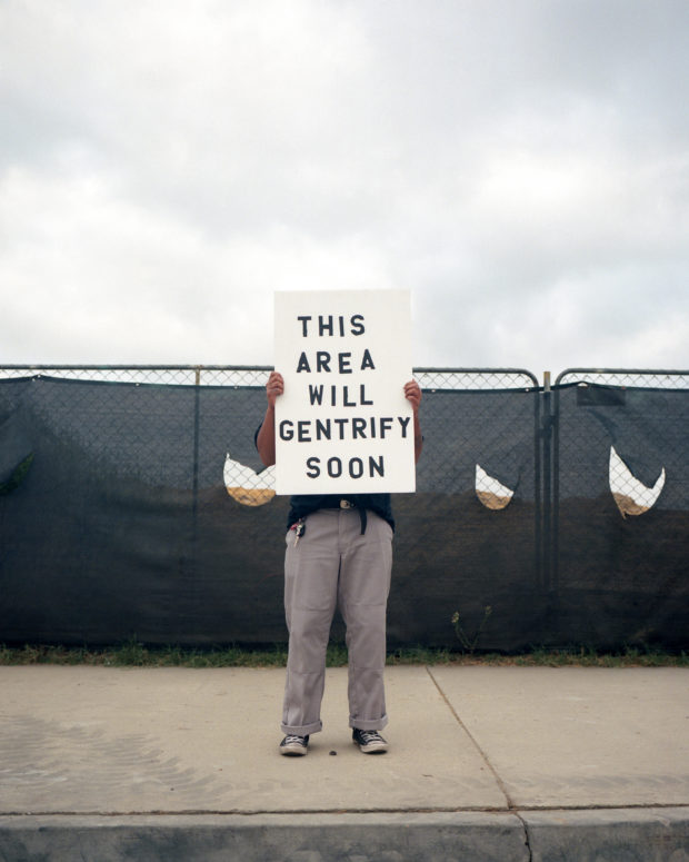 Artist stands on sidewalk in front of a material covered chain link fence holding a sign in front of his body that read "THIS AREA WILL GENTRIFY SOON" 