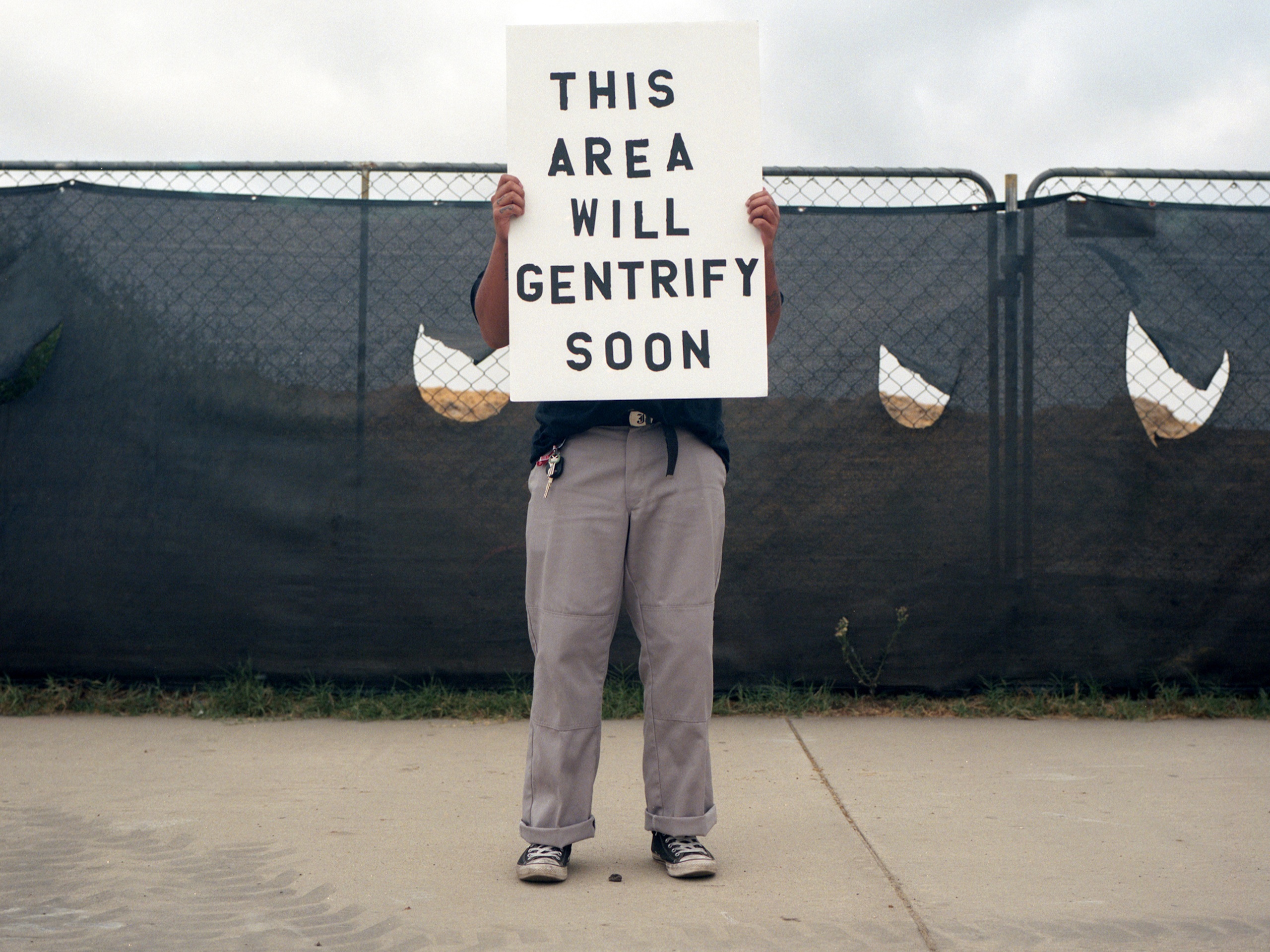 Artist stands on sidewalk in front of a material covered chain link fence holding a sign in front of his body that read "THIS AREA WILL GENTRIFY SOON"