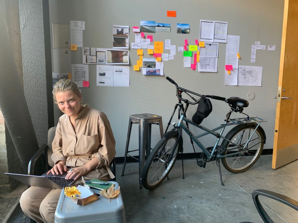 artist Ann Mirjam Vaikla sitting at a small table with artwork hanging on wall behind her and bicycle staring below the artwork