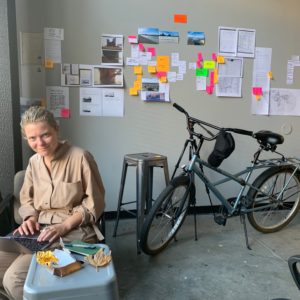 artist Ann Mirjam Vaikla sitting at a small table with artwork hanging on wall behind her and bicycle staring below the artwork